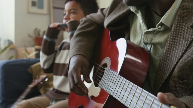 Middle aged Black man enjoying playing guitar with young grandson accompanying on harmonica, family connecting through performing music together in living room