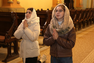 Two women kneel in church, hands folded in prayer.