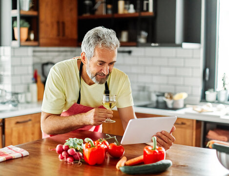 Portrait of happy senior mid aged mature man prepering meal with fresh vegatebles and following internet instructions for a recipe on a tablet computer or looking at video or website app and drinkin
