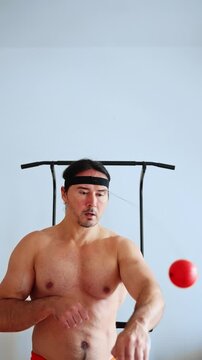 shirtless male practicing reflex ball drill, intense home workout with red tethered ball, headband, controlled punches, focused gaze, fast hand-eye coordination, sweat on torso, minimal gym frame