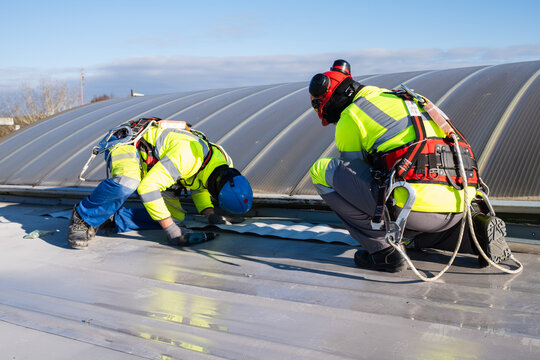 Two roofers wearing helmets and harnesses are carrying out sealing work on a flat roof near a skylight.