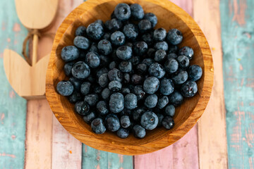 A wooden bowl filled with ripe blueberries sits on a pastel-painted rustic table. Soft natural light highlights the fresh berries, making the scene perfect for food, organic lifestyle, recipes, and he