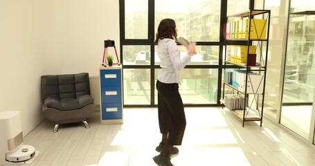 Woman dances in bright office room while waving fan of cash. Energetic dancer spins lightly shaking banknotes around smiling face after salary day