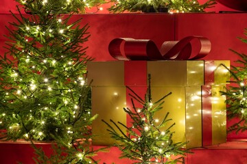 A large golden gift box with a red ribbon sits among small decorated Christmas trees with glowing fairy lights