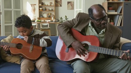 Senior African American man enjoying playing acoustic guitar with little grandson accompanying granddad on harmonica while spending leisure time together at home - Powered by Adobe