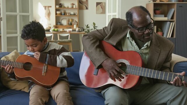 Senior African American man enjoying playing acoustic guitar with little grandson accompanying granddad on harmonica while spending leisure time together at home