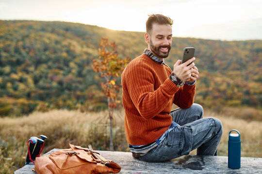 Hiking man enjoys nature using navigation app on her mobile phone and taking a break with a cup of hot tea or water bottle in a lush forest during a sunny day