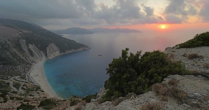 Breathtaking panorama of iconic Myrtos beach in the Greek island Kefalonia, with turquoise waters of Ionian Sea surrounded by steep cliffs, under dramatic sunset red sky, stunning Mediterranean view