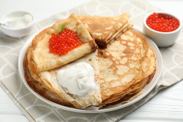 Tasty crepes with red caviar and sour cream on white wooden table, closeup