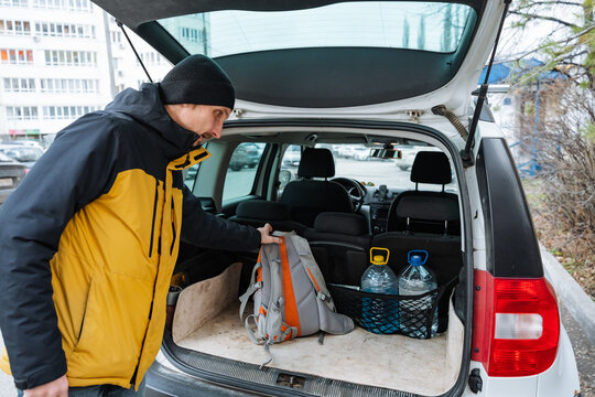 caucasian man organizing backpack and water in trunk before hike, arranging bottles and camping gear in open cargo space, autumn air and apartment backdrop, focused outdoor prep