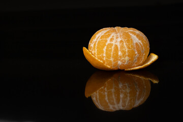 Minimalist close-up of a peeled mandarin resting on its peel with a clean reflection on a glossy black surface and with empty space.