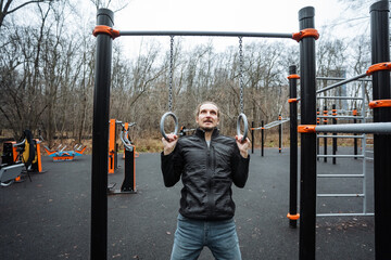 man practicing strength training in park, focused individual performing gymnastic ring exercises outdoors, man engaged in disciplined calisthenics using rings in urban park setting