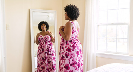 Happy plus size woman looking at reflection in mirror. Confident African American female adjusting pink floral dress in bedroom. Self love and acceptance