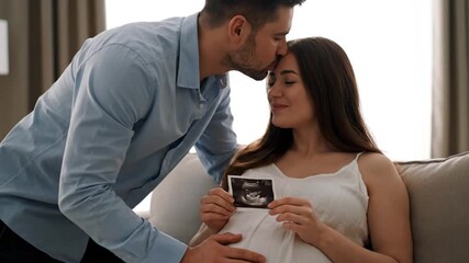 Loving husband kisses pregnant wife, happy couple holding baby ultrasound scan