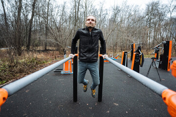 man performing controlled dips on parallel bars in outdoor fitness area, steady tempo and focused breathing, coldseason attire, gritty urban training atmosphere with bare foliage