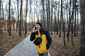 man walking through wooded path making phone call, crossbody bag and yellow jacket visible, steady stride, background of tall trees and paved trail, onthe-go communication mood