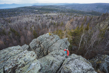 Summit quest underway, Climber scales rugged terrain confidently, Adventurous trek through forested mountain challenges, Vigorous ascent over craggy ridge amidst lush valleys and light