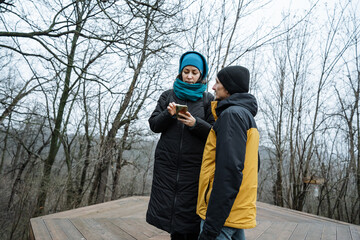 two individuals review images outdoors, two persons scrutinize pictures on forest platform, duo studies images on wooden surface amid leafless trees and cloudy surroundings