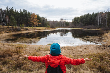 Youngster in colorful attire releasing feelings outside, Child dressed vividly stretching near shimmering water amidst nature, Juvenile in vibrant clothing reaching out at reflective pond
