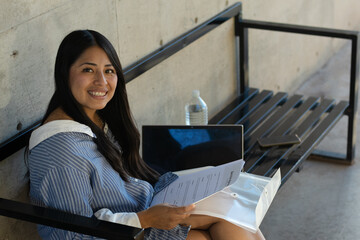 Young woman studying and working remotely outdoors on a modern bench, smiling at the camera while...