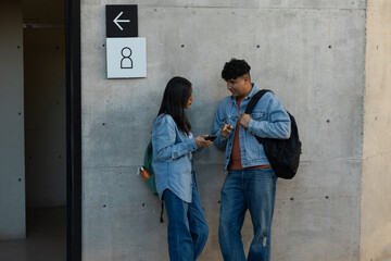 Students leaning against a concrete wall, talking and sharing content on a smartphone, representing...