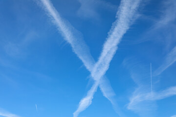 White jet contrails cross in a bold “X” against a deep blue sky on a clear day, illustrating aviation, flight paths, atmospheric science, and modern air traffic.
