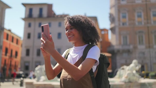 Happy female traveler with backpack recording moment beside a classic Rome Italy fountain with bright daylight, soft spray and warm holiday vibes in a lively open square.