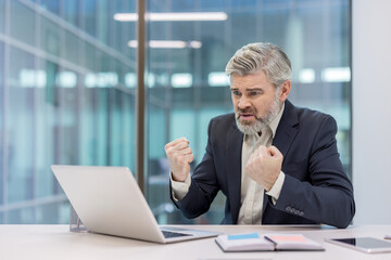 Senior businessman at a modern office desk clenches his fists in frustration and anger while staring at a laptop, showing stress, burnout and workplace despair during a crisis