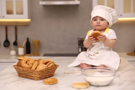 Cute baby with bun wearing chef uniform at light marble table in kitchen