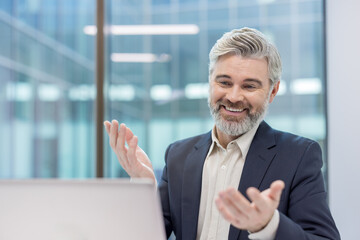 Grey-haired businessman smiling and gesturing during a virtual meeting, actively engaging in remote...