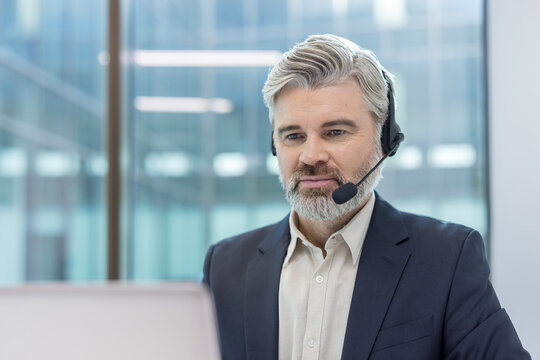 Mature man with a grey beard wearing a headset and suit jacket, working in a modern office environment, providing online customer service and technical support