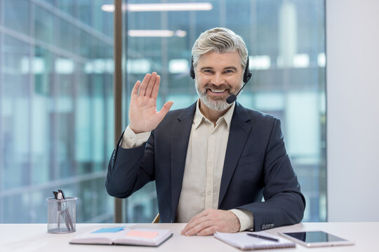 Mature businessman in suit and headset sitting at desk, smiling and waving during a virtual meeting, engaging in online coaching, remote work and professional video conferencing - Powered by Adobe