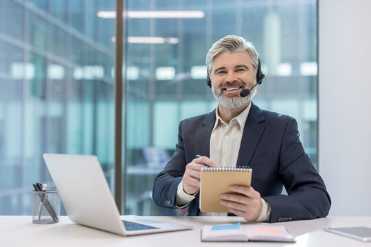 Senior professional man in a headset and suit at a modern office desk with laptop, smiling and writing in a notebook while providing friendly, reliable customer support and service