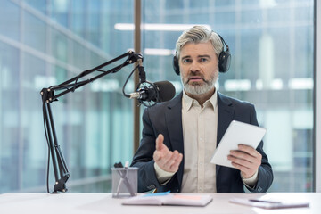 Mature man with headphones and tablet actively recording a podcast or radio broadcast in a modern studio setting, discussing current topics and sharing information