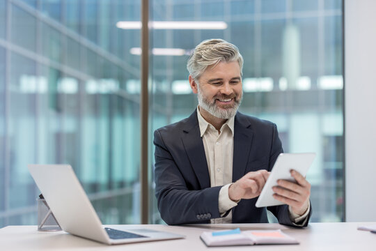 Happy mature businessman sitting at a modern office desk, smiling while using a digital tablet for work, with a laptop and notebook nearby against a blurred corporate building background - Powered by Adobe