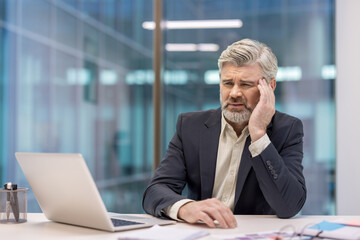Senior businessman feeling stressed and having a headache while trying to work on his laptop in a modern corporate office environment, symbolizing burnout and exhaustion