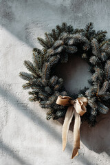 Christmas wreath made of fir branches with a decorative ribbon on a light wall in natural sunlight