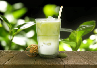 Iced matcha latte in glass, spoon with tea powder and whisk on wooden table against green leaves