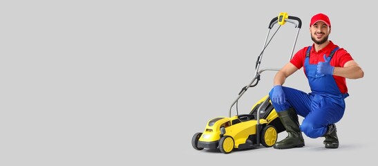 Young male gardener with modern lawn mower showing thumb-up on light background with space for text