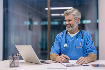 Mature doctor smiling while telecommuting from a modern office, wearing blue scrubs and a stethoscope, interacting with patients online using a laptop