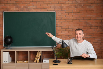 Male teacher with clipboard sitting at table in classroom and conducting lesson