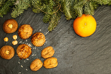 Overhead shot of Christmas baking treats including coconut cookies and walnuts on a black slate background decorated with fir branches and a fresh orange