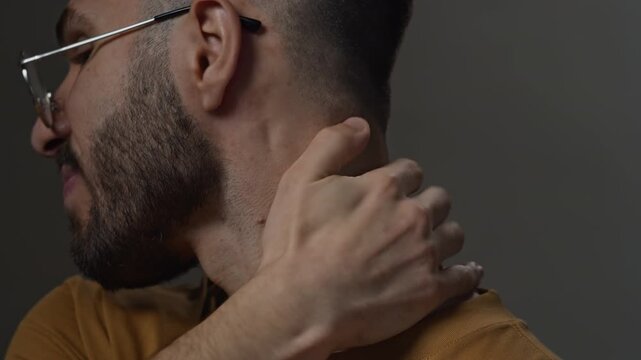 Side view close up shot of troubled young Middle Eastern man in eyeglasses scratching neck with anxiety against grey background