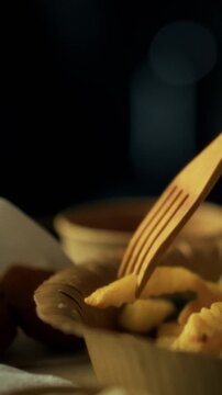 A hand carefully lifts crispy fries from a bowl using a wooden fork. The warm atmosphere suggests a relaxed evening meal, perfect for enjoying comfort food with friends or family
