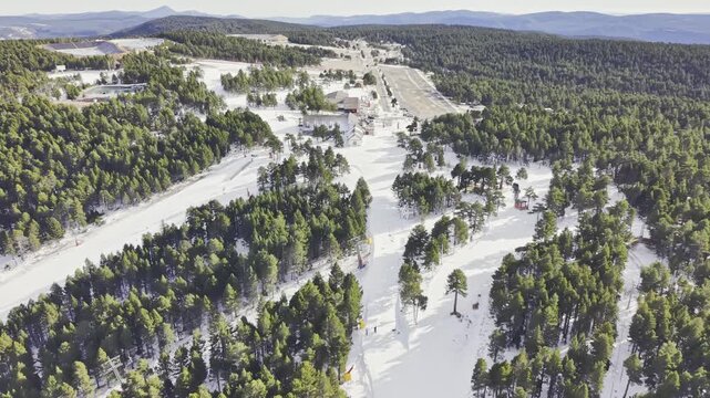 Aerial footage of a drone slowly pulling away from a wide snowy ski slope framed by evergreen pine forest, revealing expansive winter mountains, ski tracks, resort area, serene natural landscape and