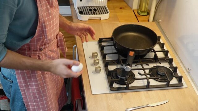 Man in apron clumsily juggling egg near gas stove awkward moment drops egg embarrassment clumsiness home kitchen cooking fail cozy evening warm light