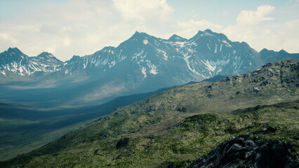 Snow capped mountains rise majestically against a clear blue sky, surrounded by lush green meadows. The sunlight casts diverse shadows on the rugged terrain, emphasizing natures grandeur.