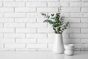 Vase with eucalyptus branches on table near white brick wall in room