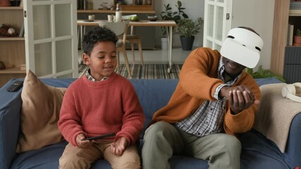 African American grandfather playing VR video game while little grandson watching and interacting nearby sitting together on couch in living room, family life scene - Powered by Adobe