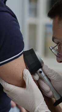 Vertical closeup shot of young Middle Eastern male doctor in white gloves using dermatoscope while examining skin irritation on forearm of unrecognizable patient in clinic